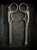 A pair of ornate metal ring scissors with decorative geometric patterns on the handles, displayed on a dark textured background.