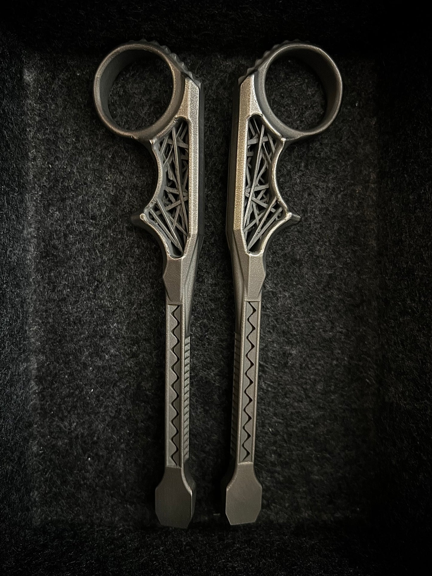 A pair of ornate metal ring tools with intricate cutout details and geometric patterns on the handles, displayed vertically on a dark textured background.