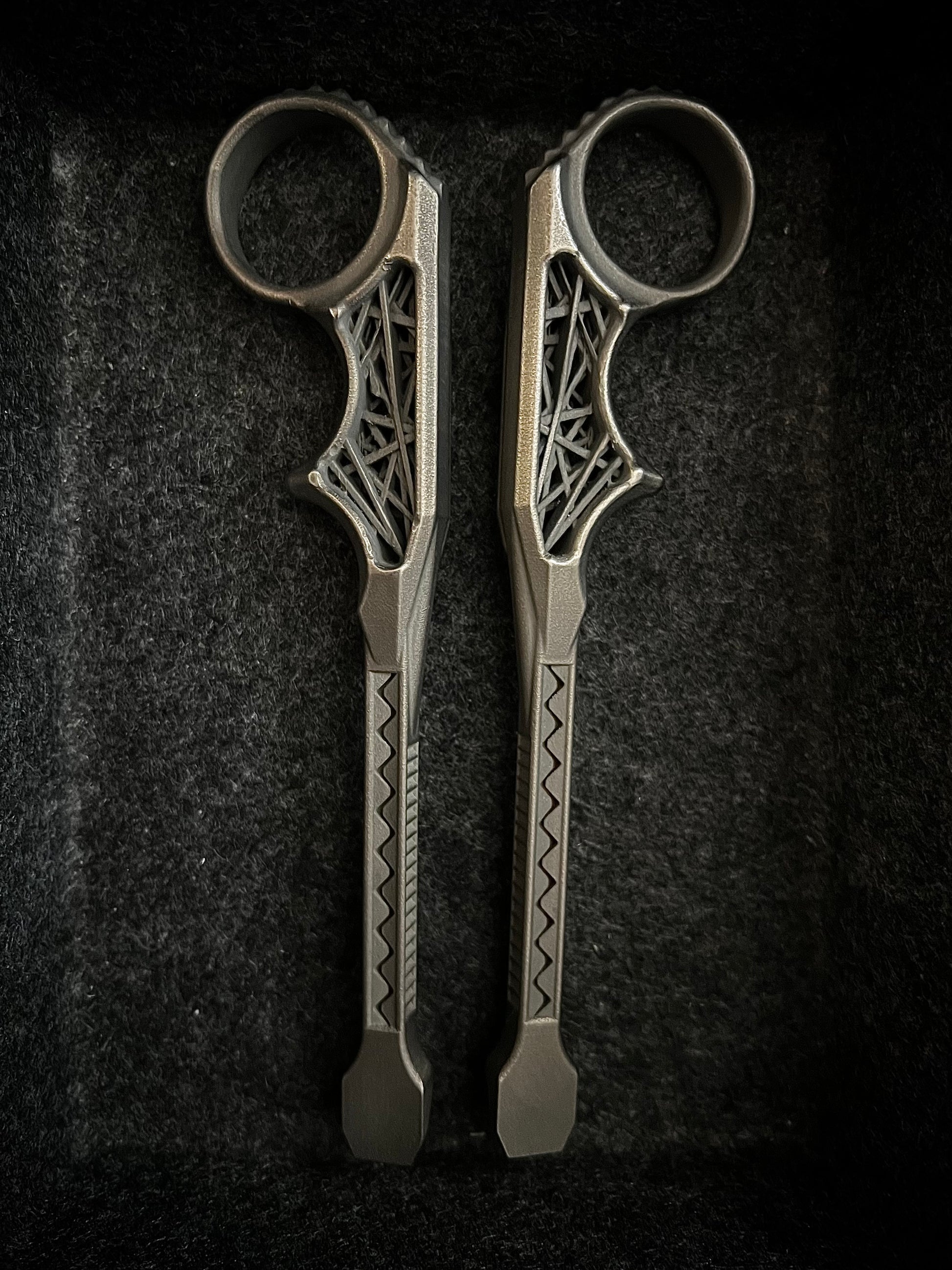 A pair of ornate metal ring tools with intricate cutout details and geometric patterns on the handles, displayed vertically on a dark textured background.