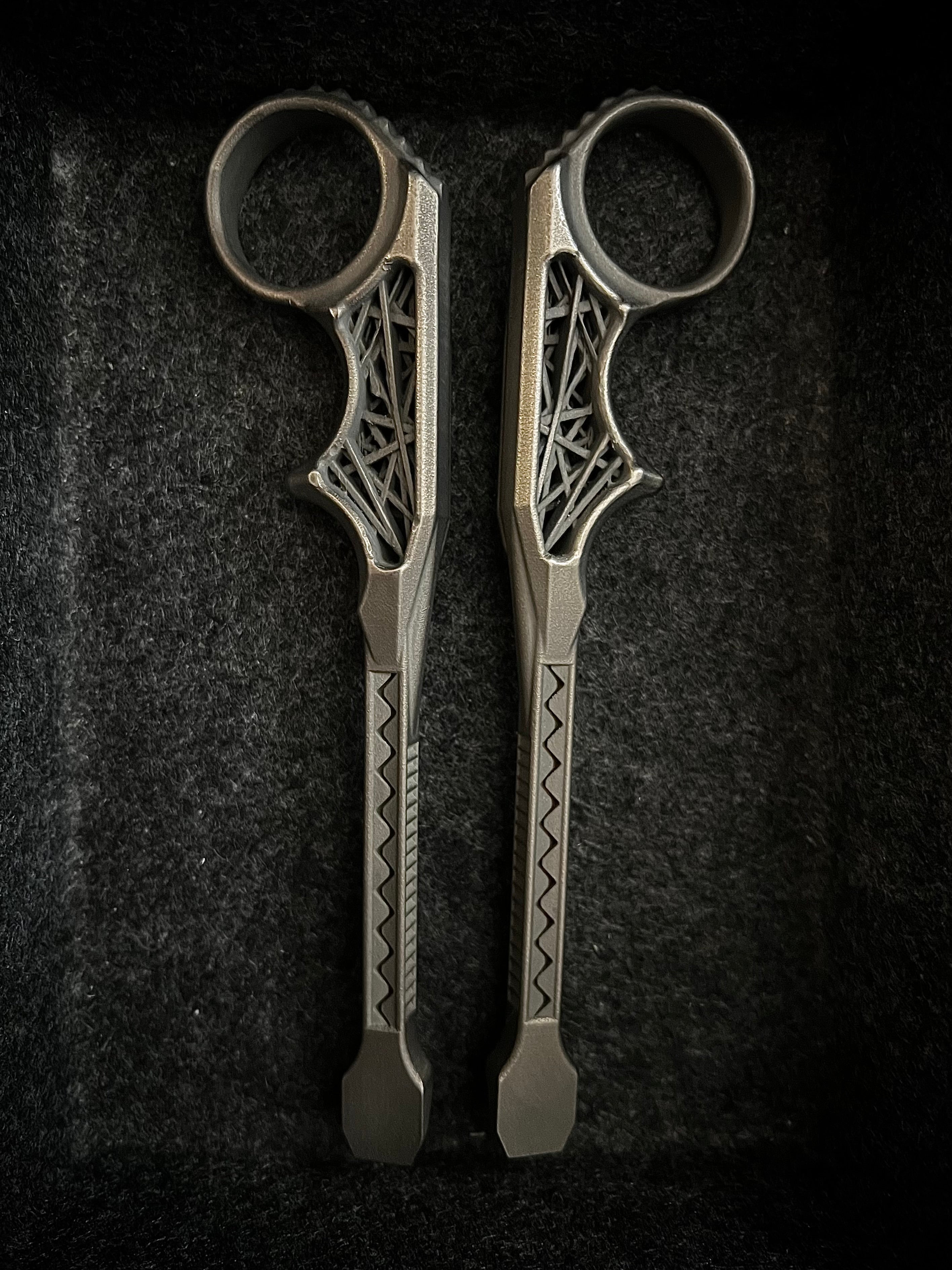 A pair of ornate metal ring tools with intricate cutout details and geometric patterns on the handles, displayed vertically on a dark textured background.