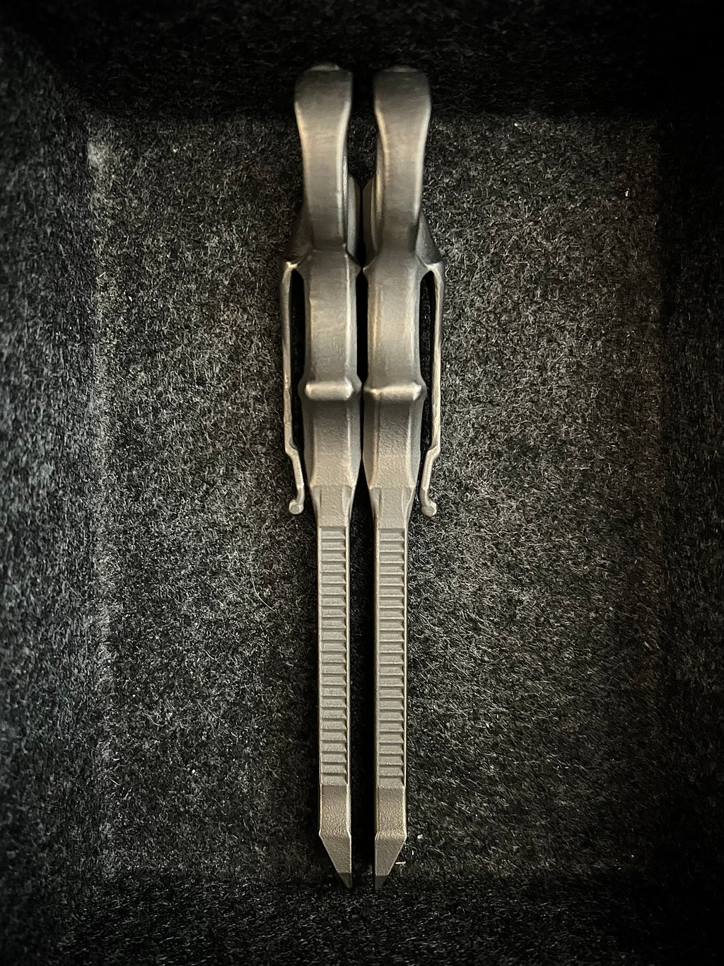A pair of metallic silver pocket knives with textured ribs on their blades, displayed vertically inside a dark gray felt-lined box.