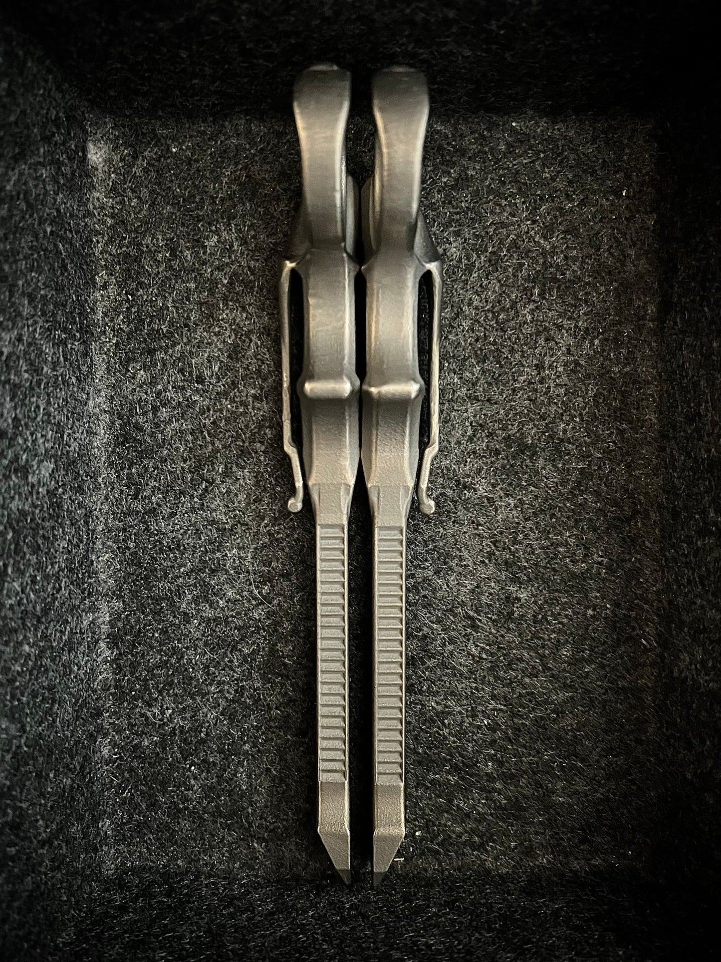 A pair of metallic silver pocket knives with textured ribs on their blades, displayed vertically inside a dark gray felt-lined box.