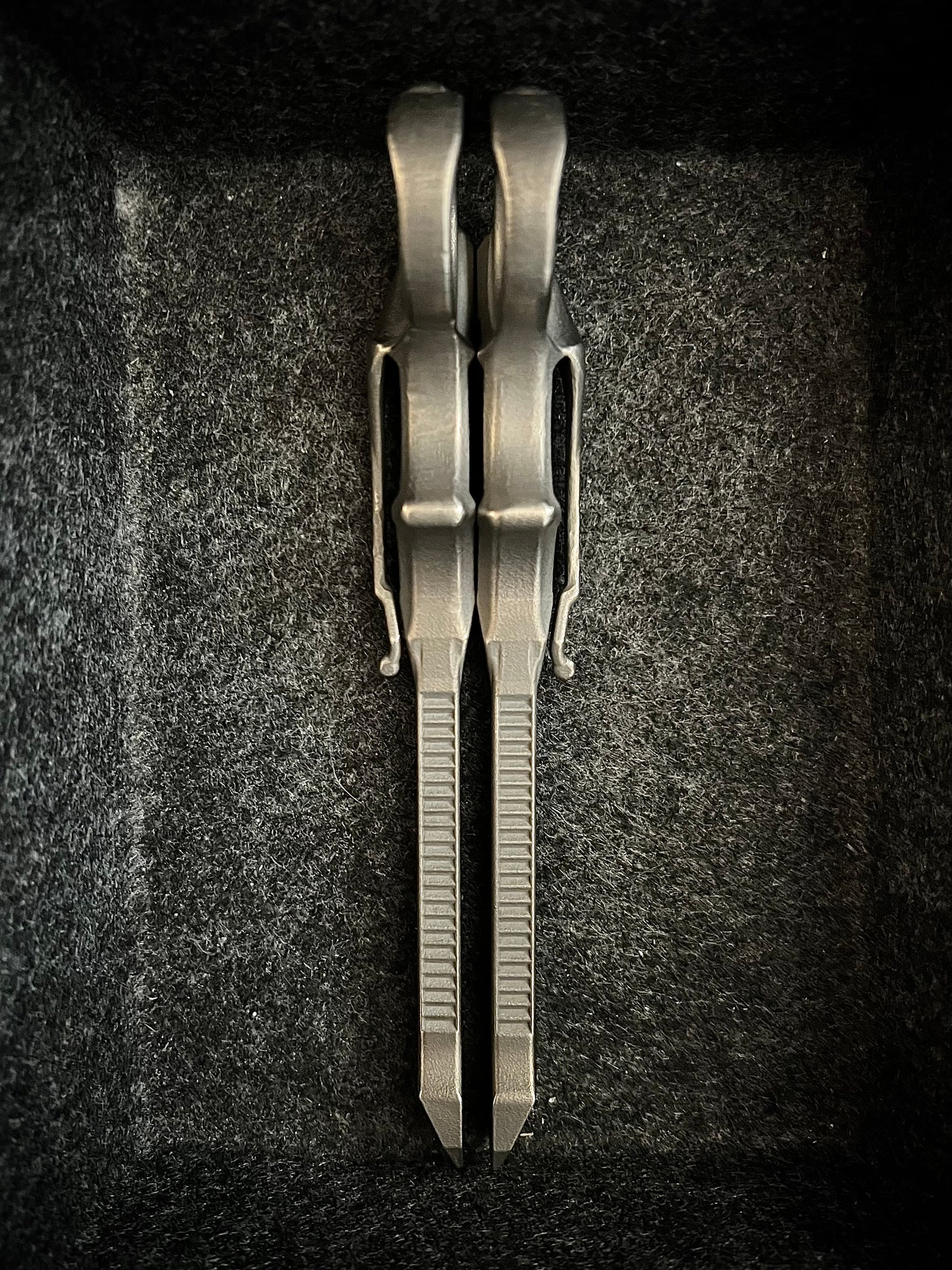 A pair of metallic silver pocket knives with textured ribs on their blades, displayed vertically inside a dark gray felt-lined box.