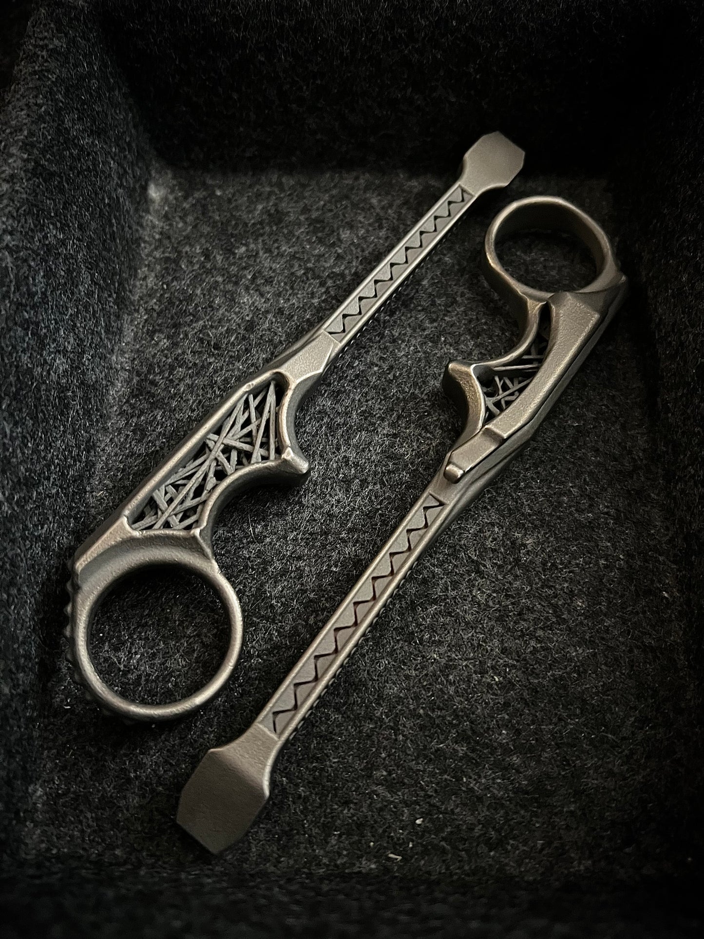 A pair of silver-toned metal finger tools with decorative geometric cutouts and textured handles, displayed inside a dark textured box.