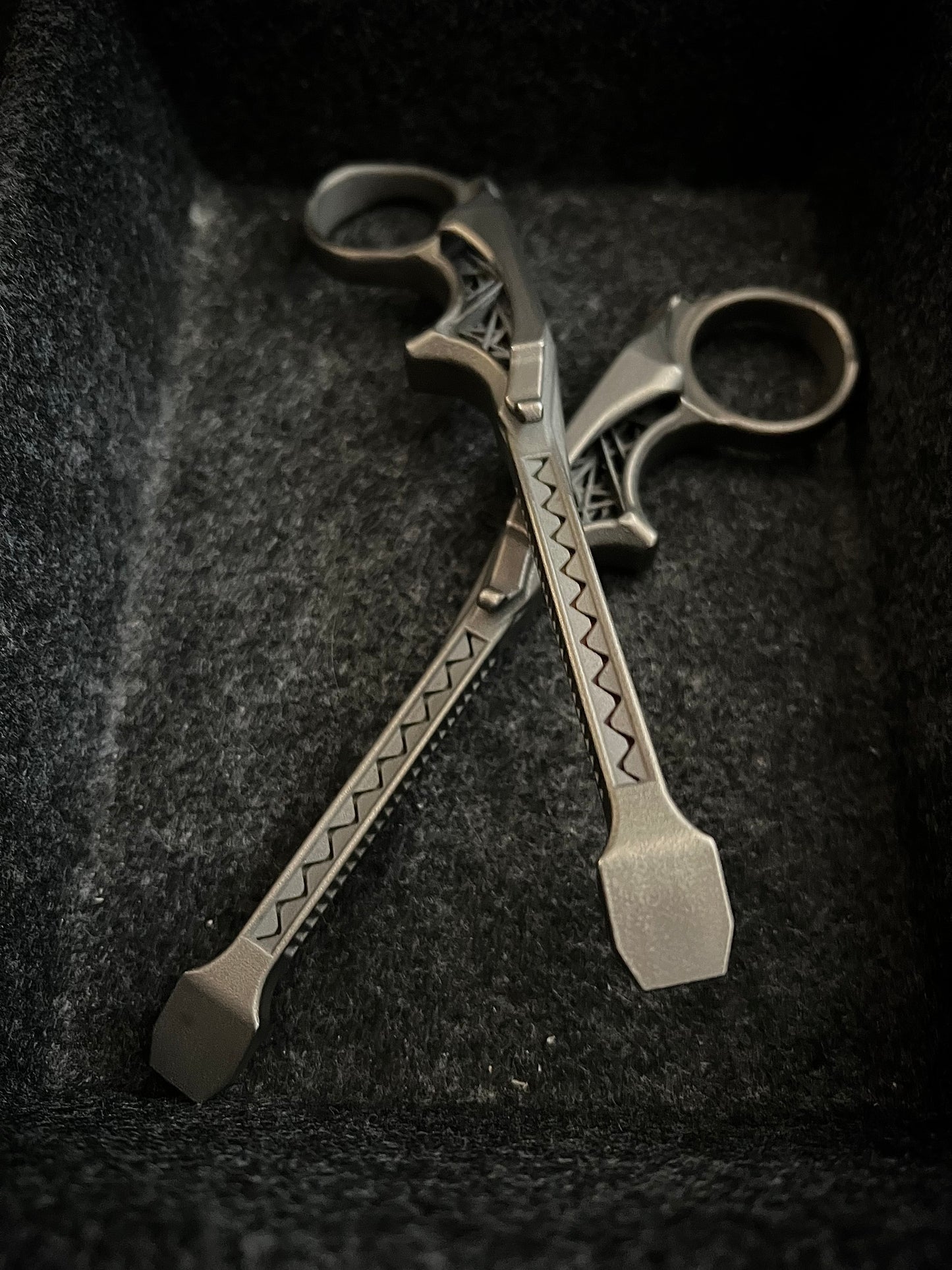 A pair of ornate silver-toned metal tools with decorative engraved patterns, resting crossed on a dark textured fabric surface.