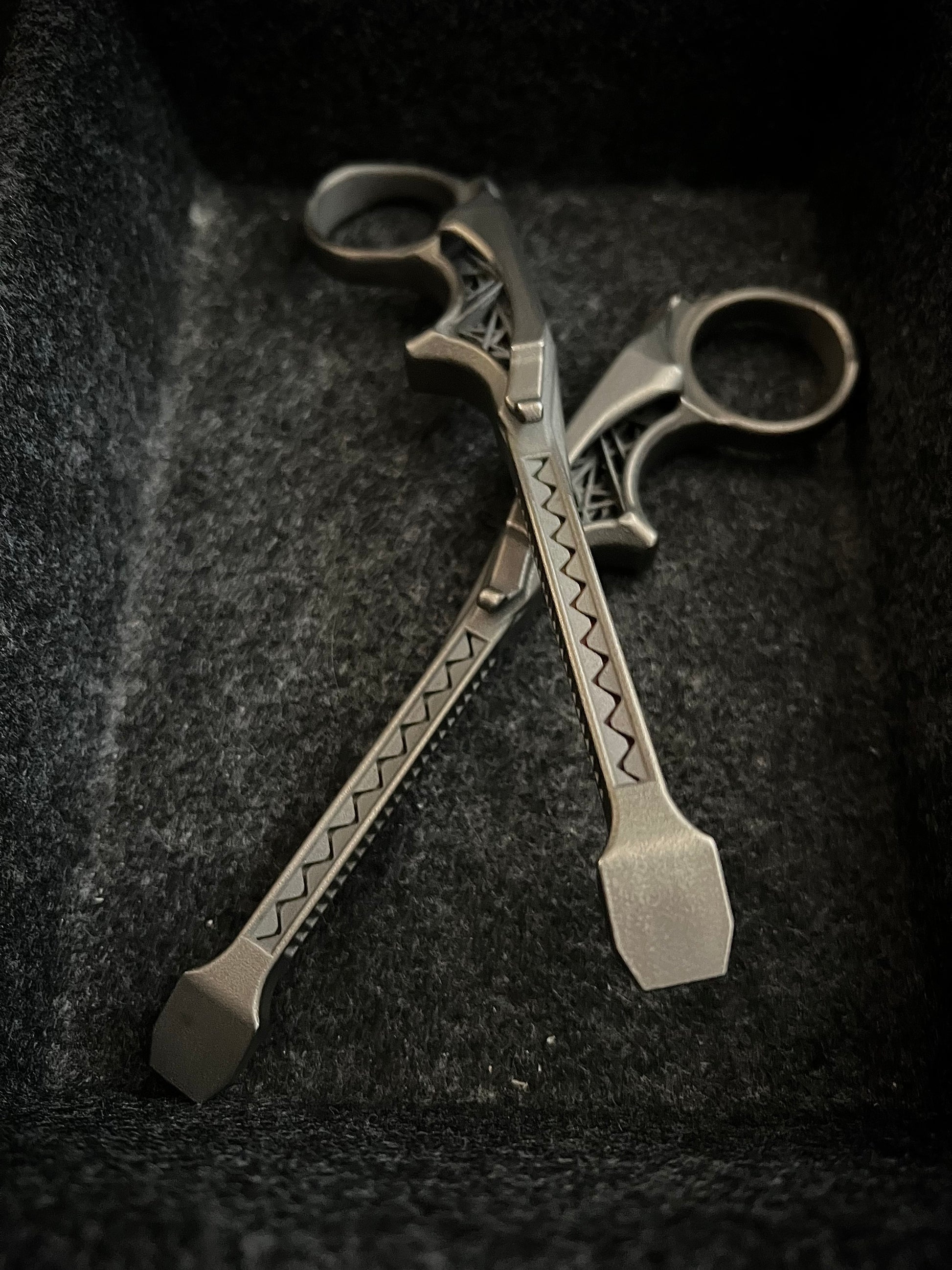 A pair of ornate silver-toned metal tools with decorative engraved patterns, resting crossed on a dark textured fabric surface.