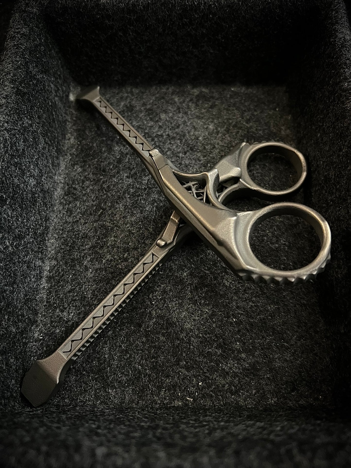 A pair of metallic scissors with textured, patterned handles resting inside a dark gray felt-lined box.